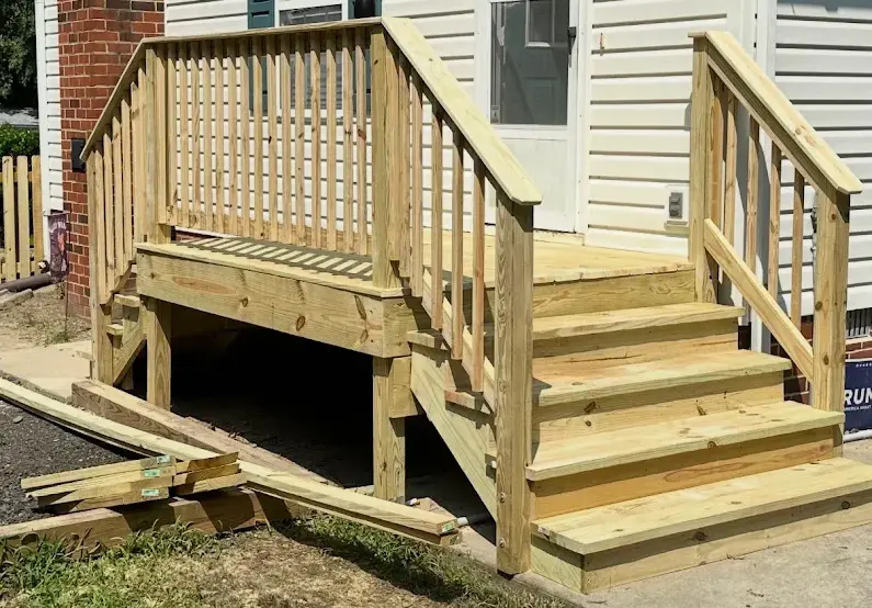 Newly constructed wooden porch deck and entry stairs with wooden railings leading up to the doorway of a house with white siding.
