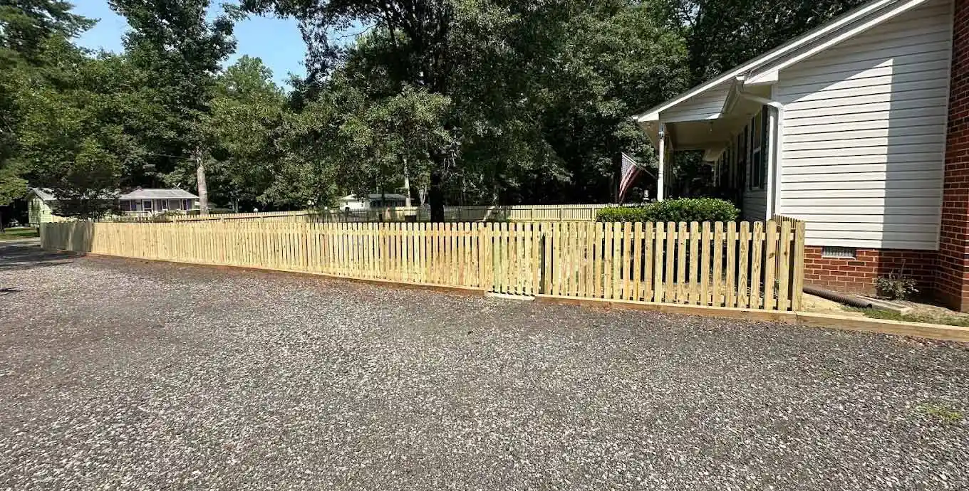 Long wooden picket fence running along a gravel driveway in front of a residential house surrounded by shade trees.