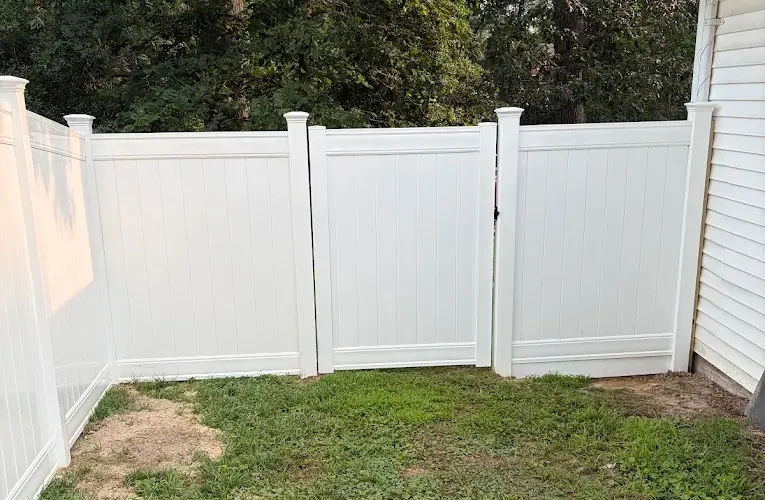 White vinyl privacy fence enclosure featuring a central swing gate on a green lawn next to a residential house.